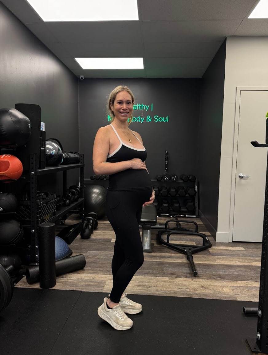 Ali, a strong mom, smiling with a hand on her pregnant belly in the studio at Balanced Health Fitness in Seaford, NY.