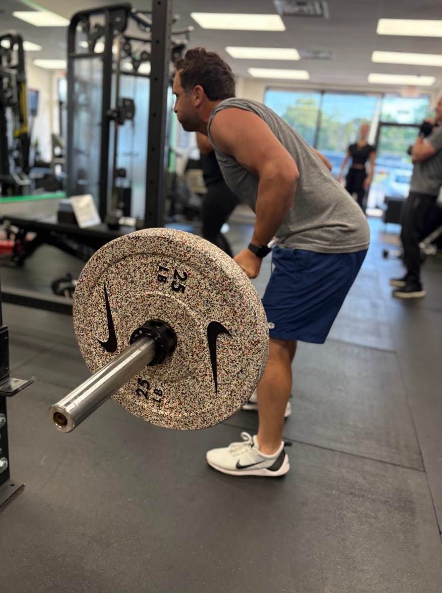 A busy dad of two, a Balanced Health Fitness member, hinged over a loaded barbell with other members training behind him.