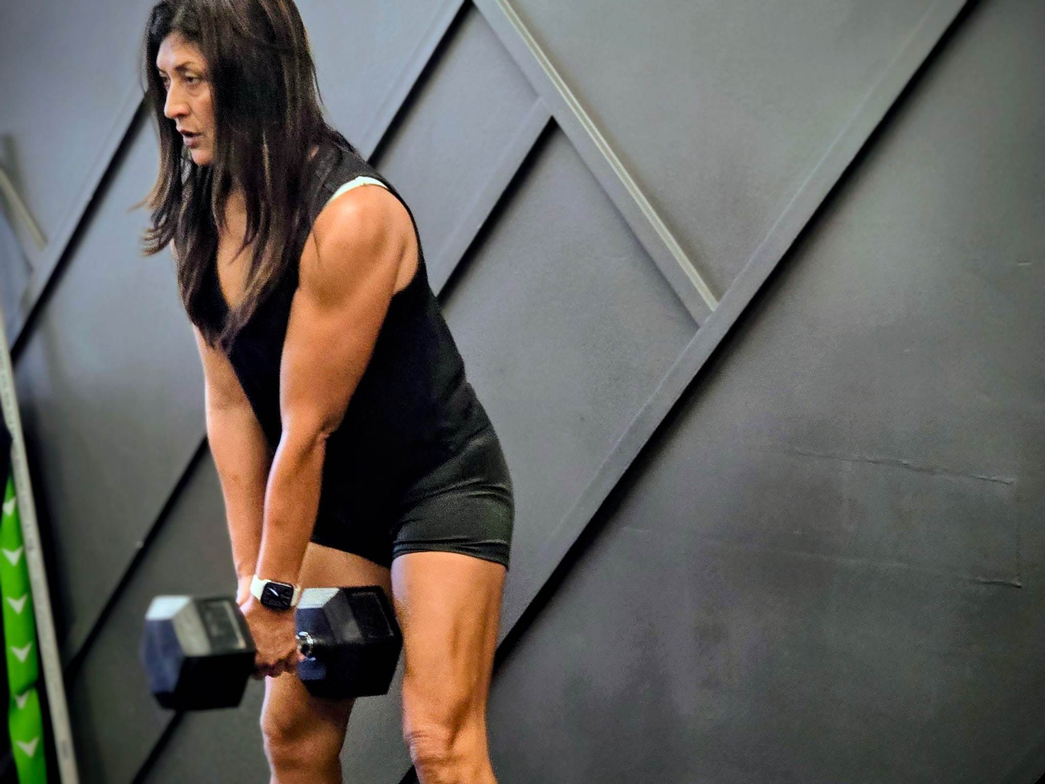 A member over 50 holding a heavy dumbbell in both hands in a hip hinge, in front of the studio's paneled wall at Balanced Health Fitness in Seaford, NY.